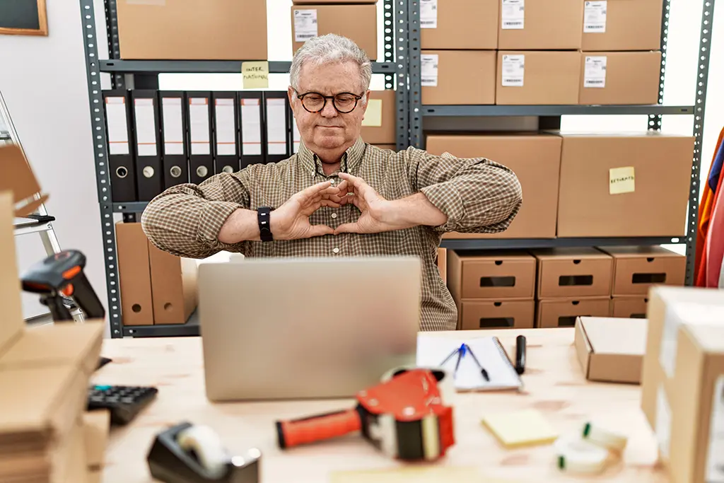 مرد میانسال در حال کار با لپ تاپ و تجارت عمده اینترنتی و علامت قلب را با دست نمایش می دهد