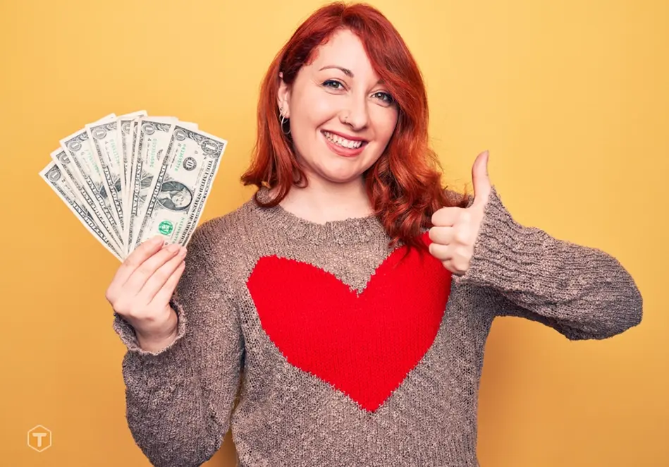 یک خانوم خندان با لباس کاموایی و طرح قلب قرمز بر روی آن و تعدادی دلار در دست راست و با دست چپ هم علامت چپ علامت لایک را نشان می دهد